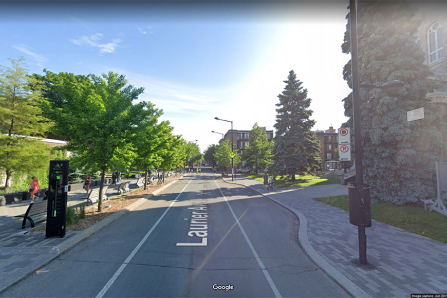 Painted bike lanes on either side of a single one-way vehicle traffic lane; pedestrians and cyclists out on a sunny day.