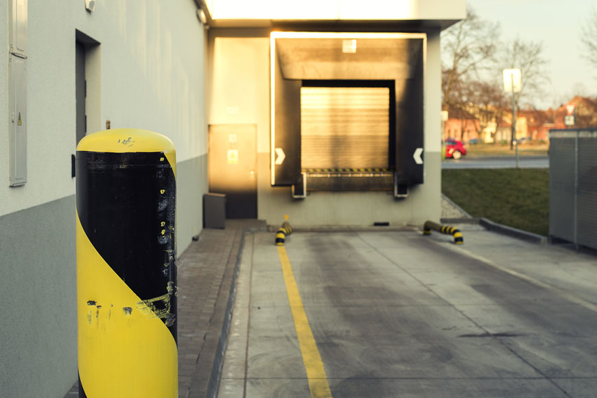 A scuffed safety bollard painted yellow and black guards the wall beside a loading dock