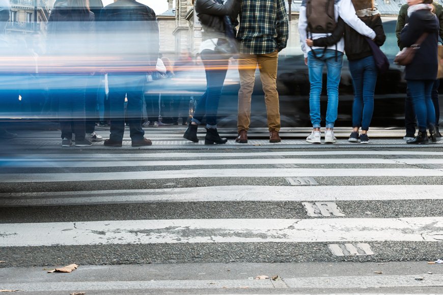 People wait at a crosswalk on a city street while cars blur past