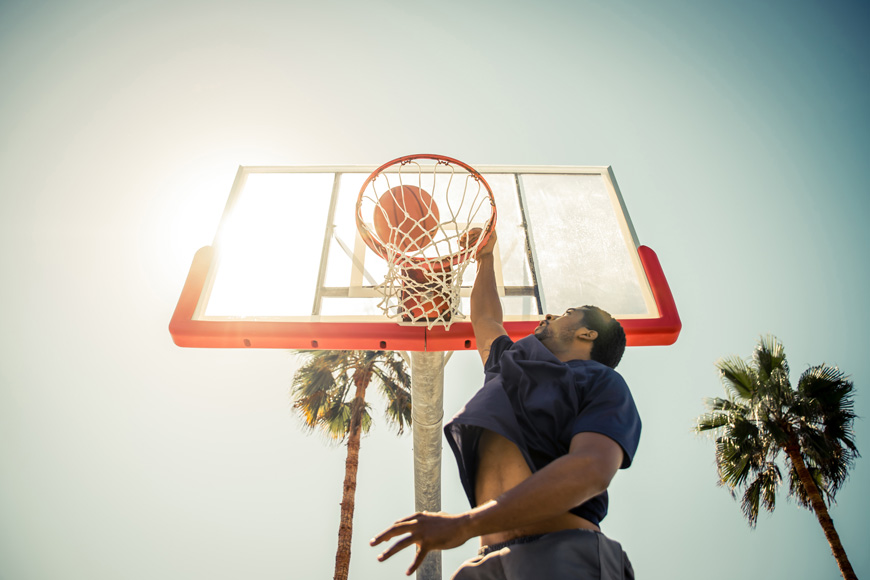 A man leaps above the camera to dunk a basketball on a sunny court surrounded by palm trees.