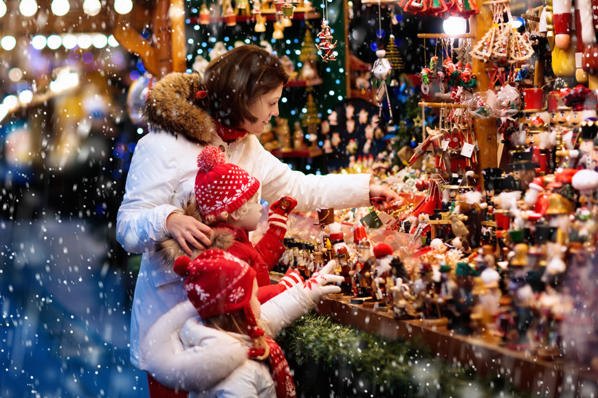 A mother and two children look at Christmas ornaments at a vendor in a snowy outdoor market.