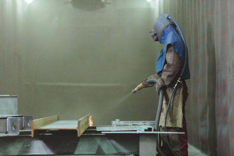 A worker covered in protective gear and mask aims a sandblaster at a piece of carbon steel