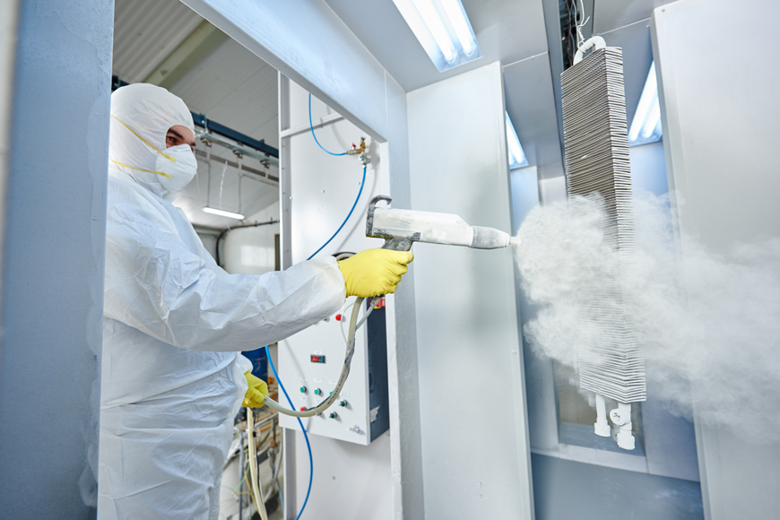 A man in coveralls and a mask shoots a stream of powder from a gun at a hanging metal item