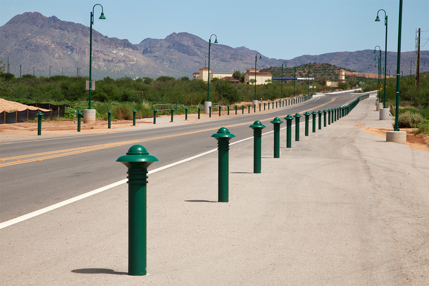 An empty road running toward rocky brown mountains is lined by scrubby bushes and green bollards.