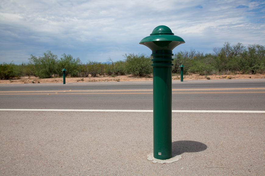 A closeup of a green bollard shows little wear. Scrub bushes and a blue sky frame the installation.