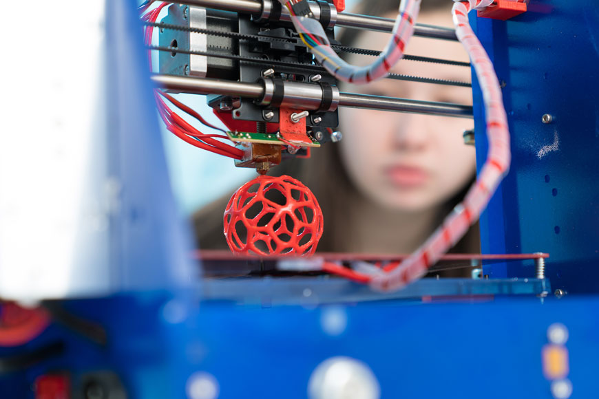 A young woman looks at a 3D printer creating a ball-shaped prototype