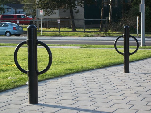 Two black outdoor bicycle racks of post and ring style stand on pavement beside a lawn