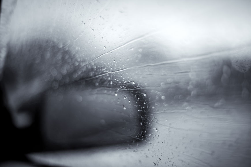 A car’s side-mirror is seen blurrily through a foggy window streaked with rain