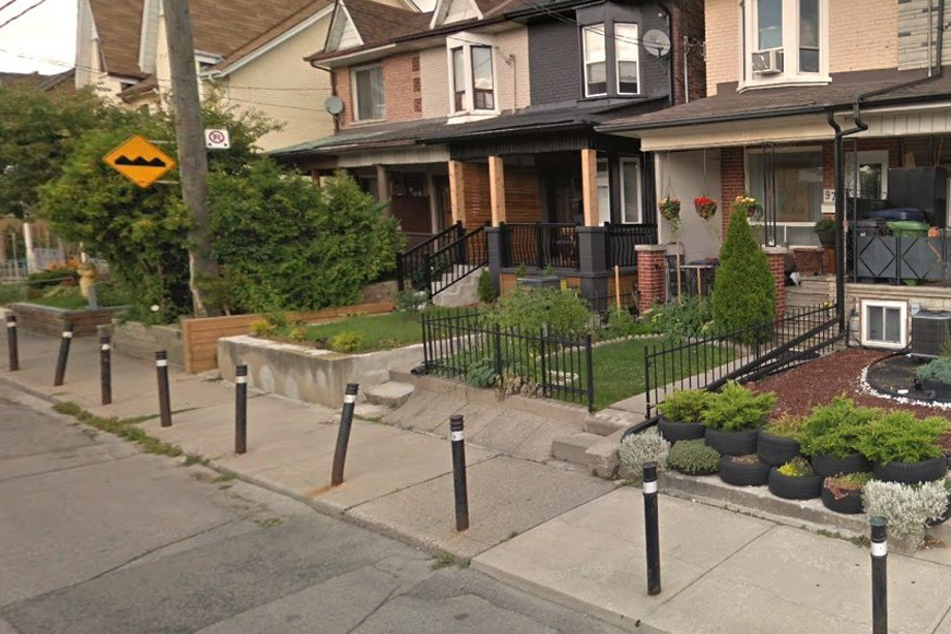 On a narrow urban sidewalk, mid-block bollards protect a line of homes