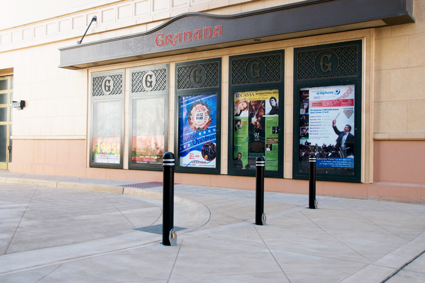 Bollards with padlocks at their base separate a sidewalk in front of posters at concert hall.