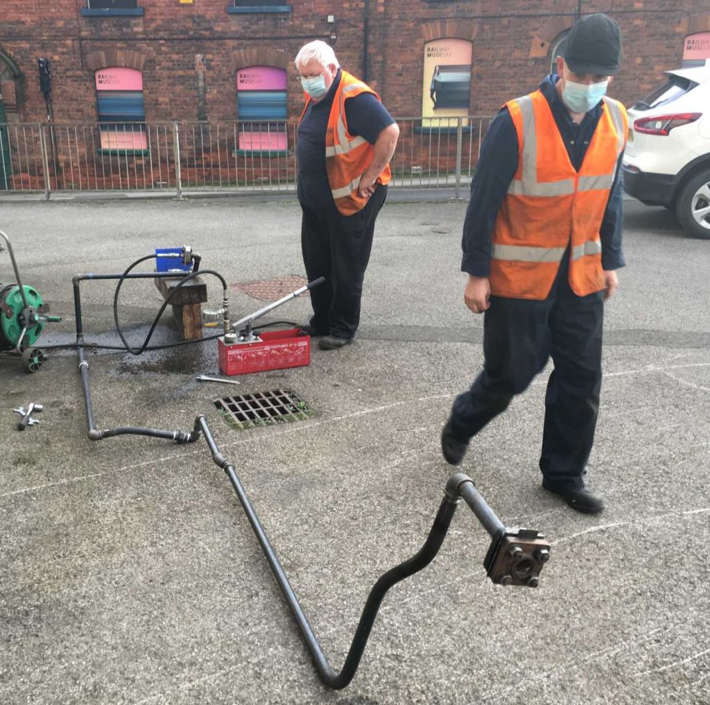 Two men in black clothes and orange hi-vis jackets looking at pressure testing equipment