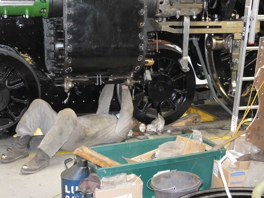 A person in grey overalls lies under a locomotive frame, reaching up to work on part of it